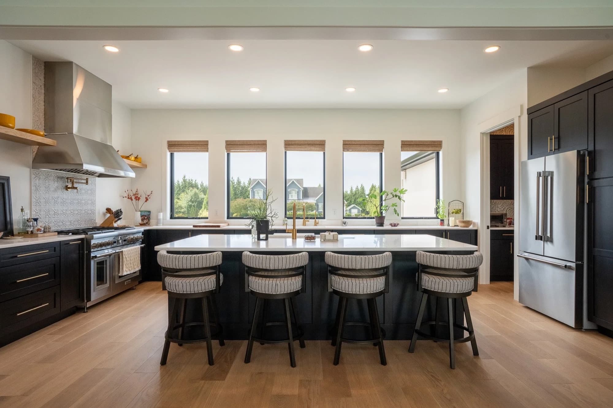 A custom kitchen with a long dark island, cane backed stools, white oak floors and a row of windows over the sink