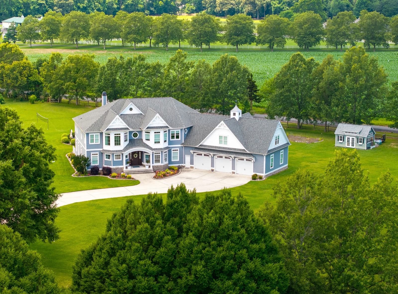 Aerial view of a blue shingled house set among mature trees beside open farmland