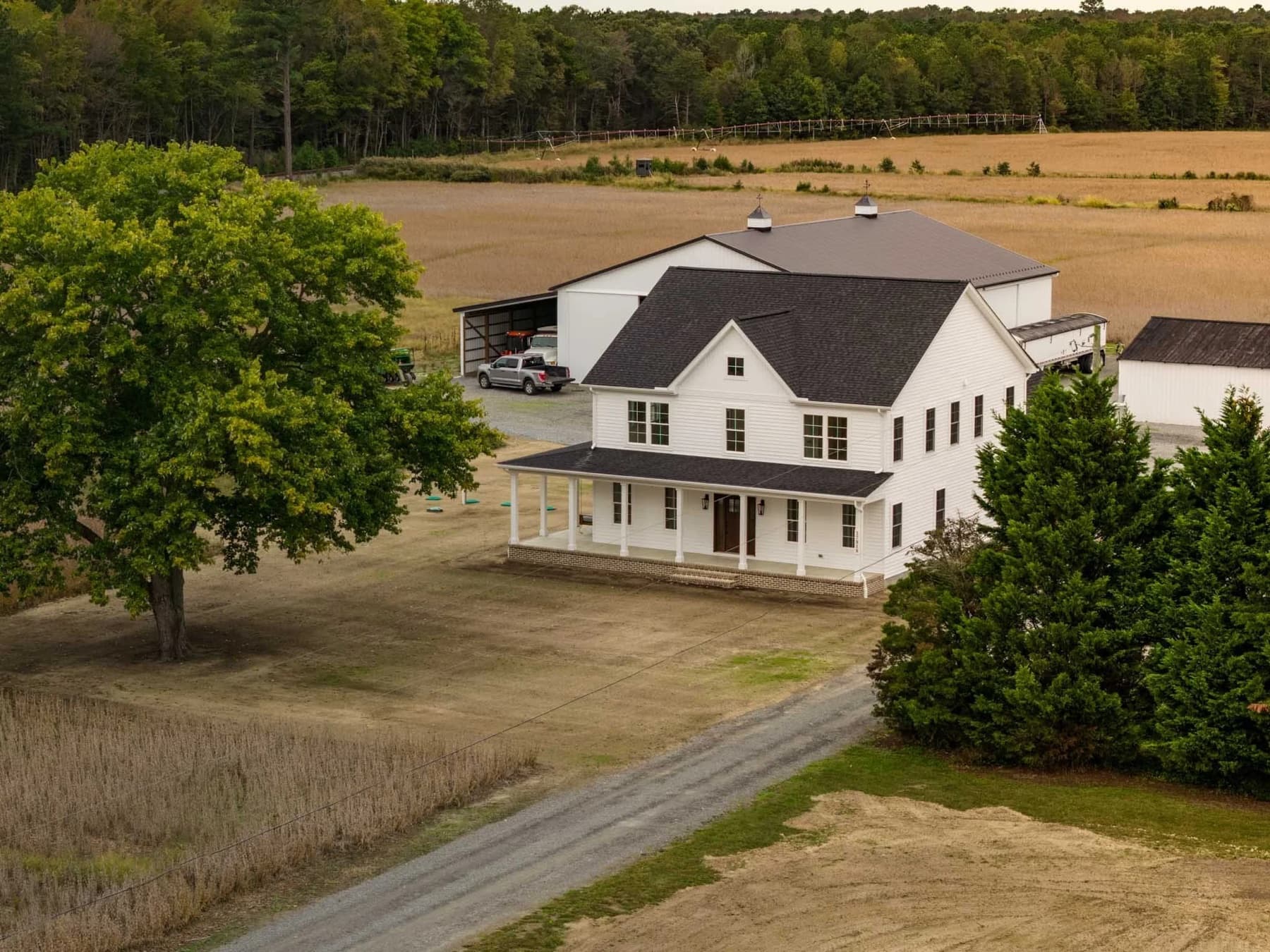 A white farmhouse and matching barn on a gravel drive at the edge of a harvested field